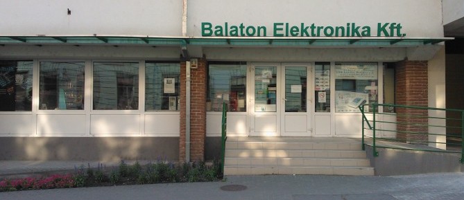 boltkep bejarat A Balaton Elektronika Kft. üzletének bejáratát mutatja. A bejárat fehér színű műanyag ajtót és hatalmas üveg felületet mutat. A cégfeltirat zöld színű betűvel van felirva az ajtó felett.